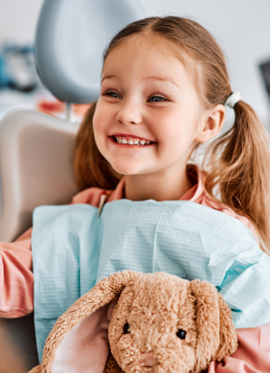 A smiling child with pigtails sits in a dental chair, holding a plush bunny. The background is a blurred dental clinic environment.