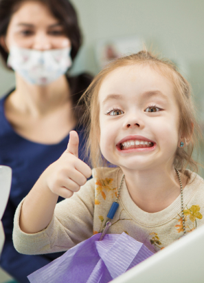 A smiling child gives a thumbs-up while sitting in a dental chair, with a masked person in the background. Indoor setting, no landmarks visible.