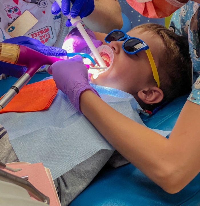 A child wearing sunglasses receives dental treatment from two people wearing gloves and medical uniforms in a bright, colorful dentist's office.