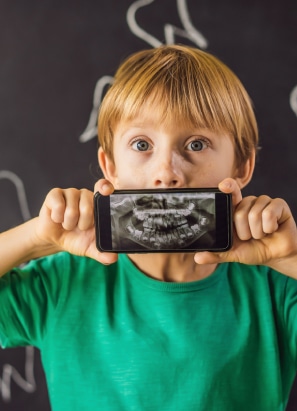 A child in a green shirt holds a smartphone displaying a dental X-ray in front of their mouth, standing against a blackboard background.