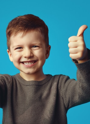 A smiling child with brown hair, wearing a gray sweater, gives two thumbs up against a plain blue background. No recognizable landmarks are visible.