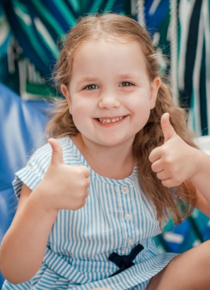 A smiling child in a striped dress gives two thumbs up, seated in front of a colorful, patterned background. No recognizable landmarks or historical buildings.