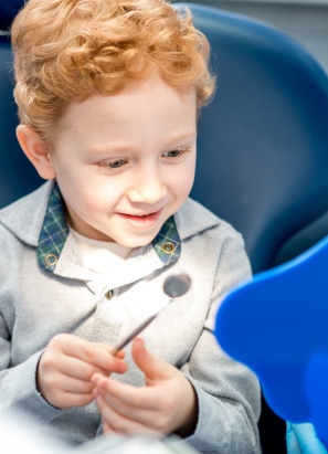 A child, seated in a dentist's chair, smiling while holding a dental mirror, with bright lighting and dental equipment visible in the foreground.