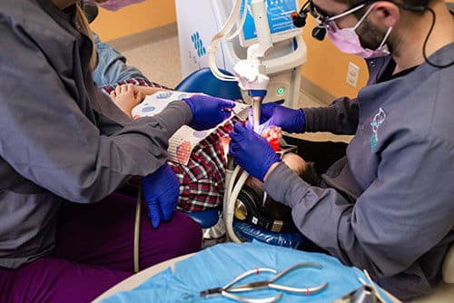 A child receives dental treatment from two masked and gloved persons in a dental office. The child is reclined in a chair.