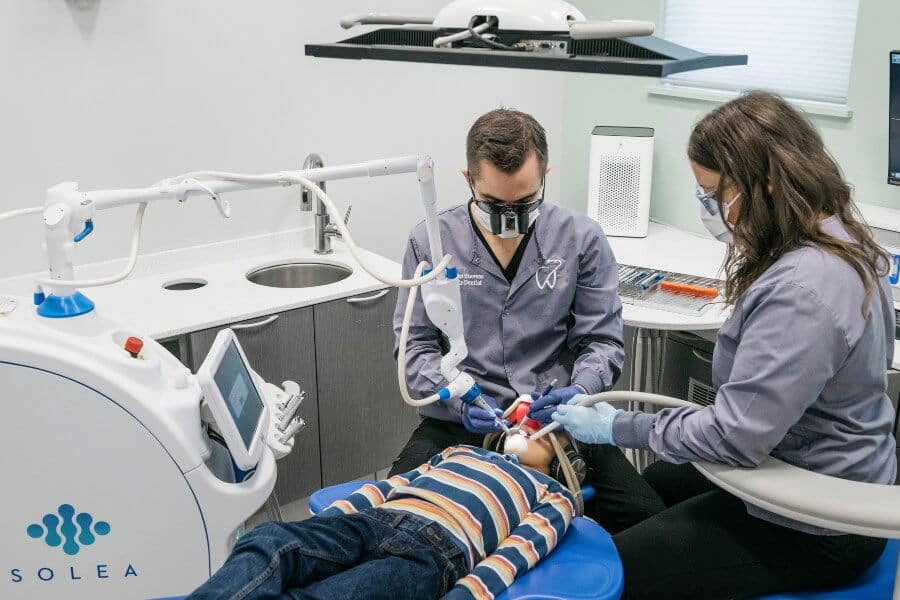 Two dental professionals treating a child in a modern dental clinic, using advanced equipment. The child is reclining in a blue dental chair.