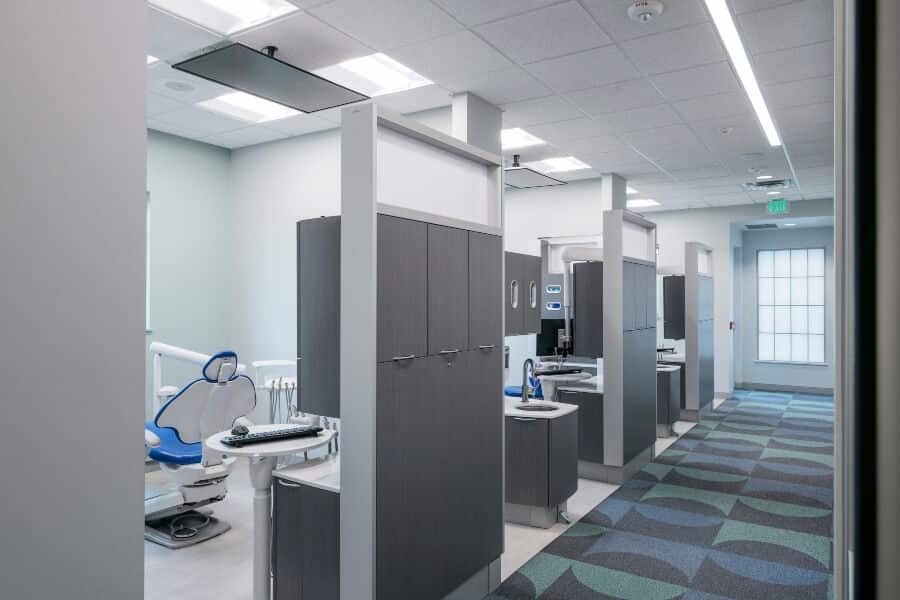 A modern dental clinic interior with multiple dental chairs, partitions, and a patterned carpet. Bright lighting and a clean, organized environment are visible.