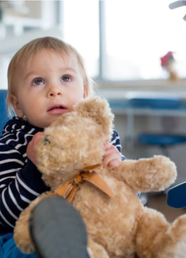 A toddler hugging a teddy bear looks upwards, seated indoors near a window with blurred background. No recognizable landmarks or historical buildings.