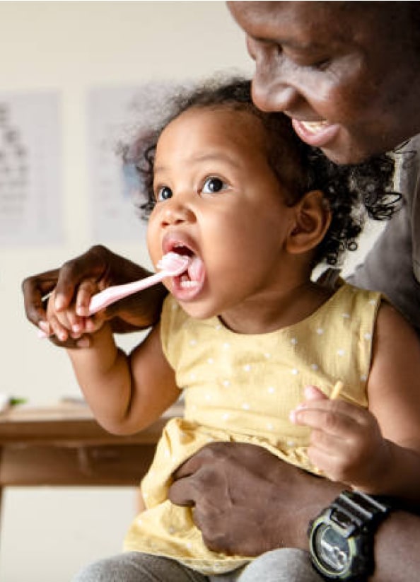 A person helping a toddler brush their teeth, with educational posters in the background indoors. The toddler is holding a toothbrush.