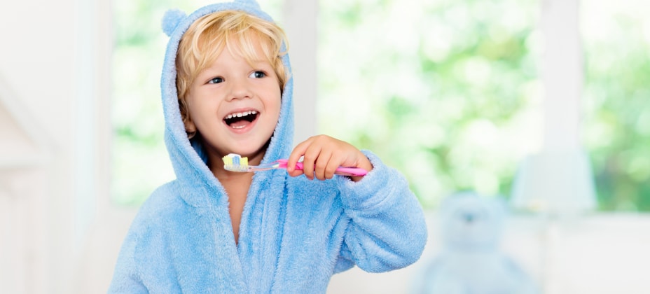 A young child in a blue bear-themed robe brushes their teeth with a pink toothbrush, smiling in a brightly lit room.