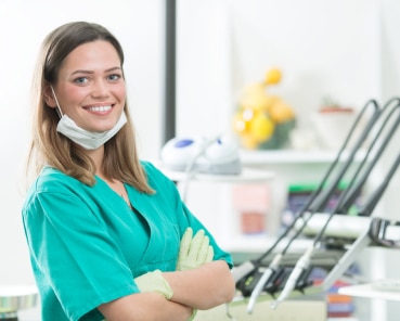 A person wearing green scrubs and a surgical mask stands smiling in a medical or dental clinic, with equipment and shelves in the background.