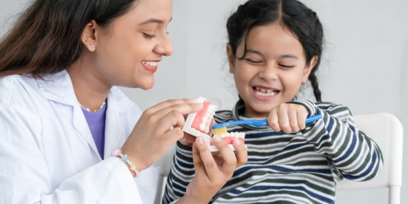 A person in a white coat teaches a child how to brush teeth using a dental model. The child smiles and holds a toothbrush.