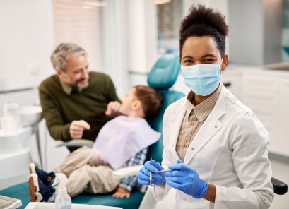 A dentist, wearing a mask and gloves, prepares dental tools while another person sits in the chair with a child in a dental clinic.