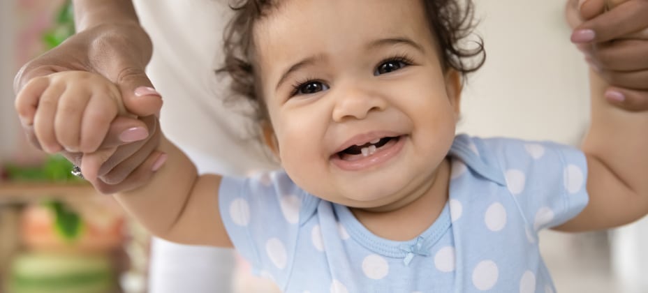 A smiling baby in a blue polka dot outfit holds hands with a person, indoors with a blurred plant in the background.