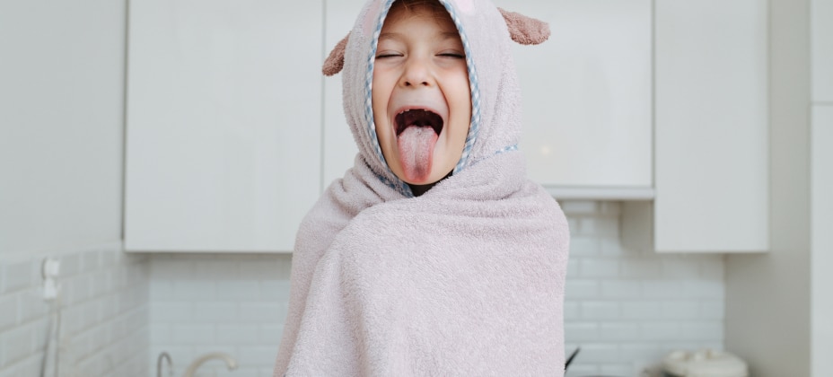 A child wrapped in a towel, playfully sticking out their tongue, stands in a modern kitchen with white cabinets and tiled backsplash.