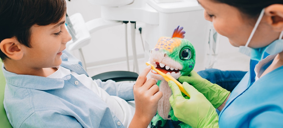 A child and a person practice tooth brushing on a colorful toy dinosaur in a clinical setting, likely a dental office.