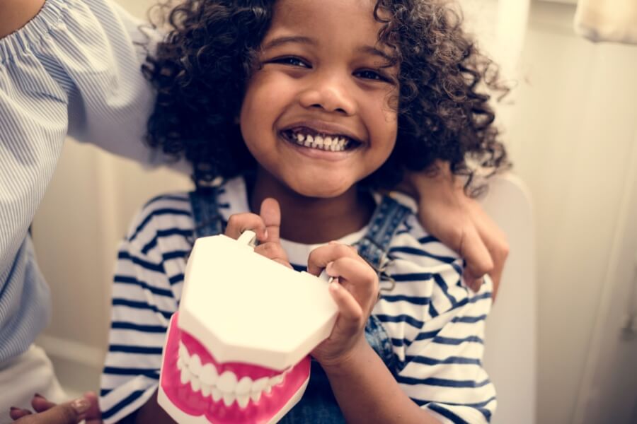 A child joyfully holds a toy set of teeth, sitting next to a person in a bright room, with a big smile.