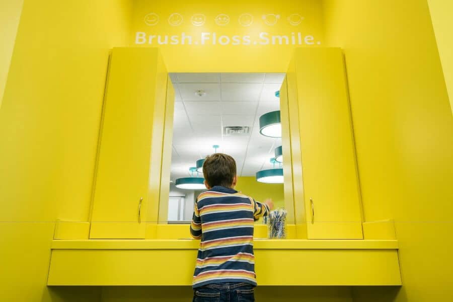 A child stands before a yellow bathroom mirror with the words "Brush. Floss. Smile." overhead, wearing a striped shirt. Bright, modern setting.