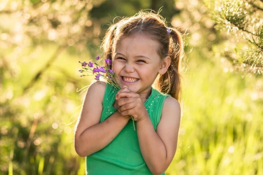 A child with ponytails smiles, holding a bouquet of purple flowers, standing outdoors in a sunlit, grassy field.