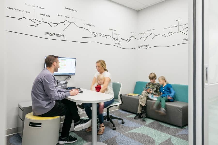 A person and child are seated at a table in a modern room, with mountain wall art, while others sit on a sofa.