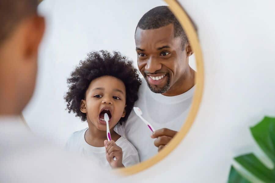 A person and child smile in a bathroom mirror while brushing their teeth. The scene is bright and cheerful, emphasizing daily routines.