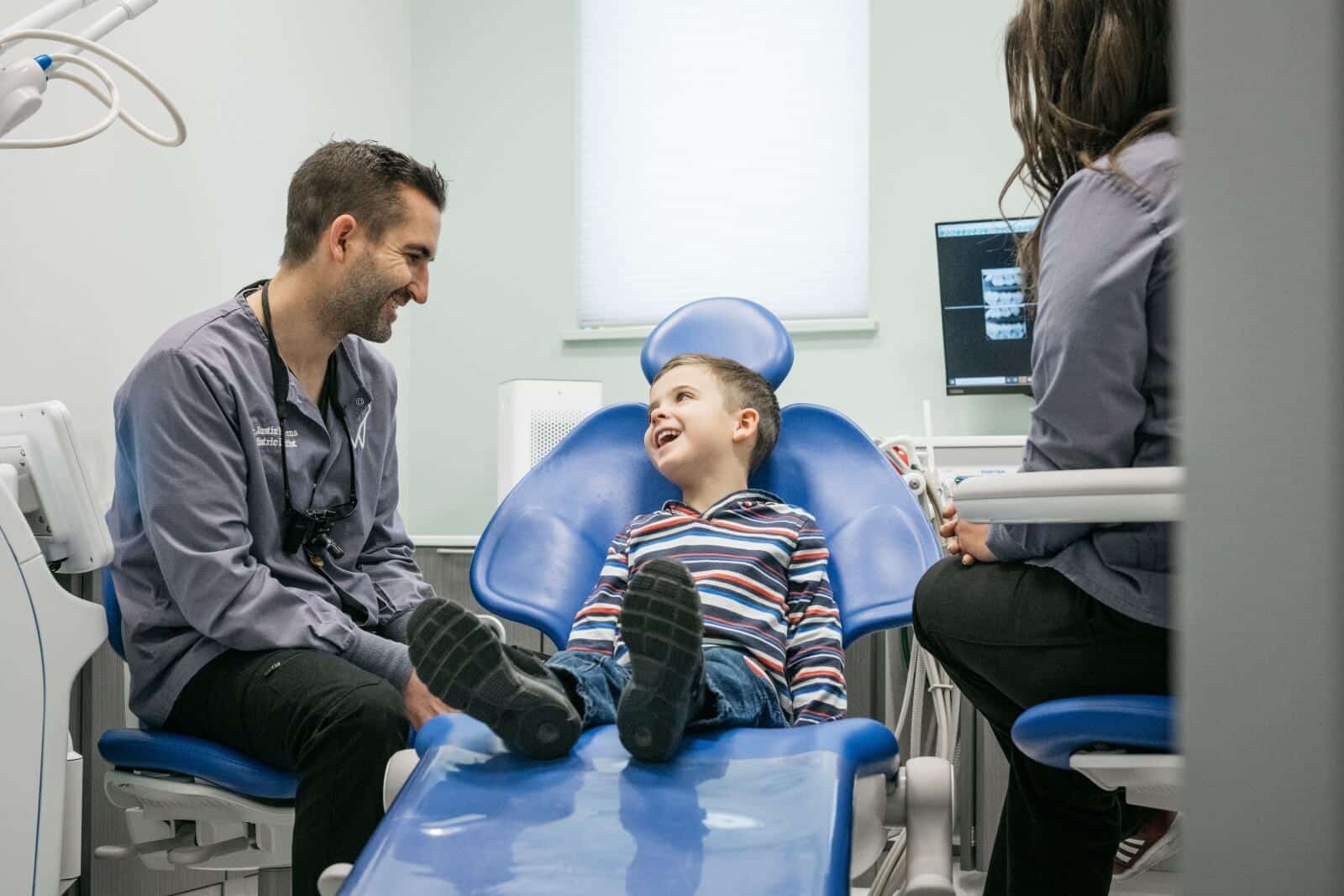 A child sits on a dentist's chair, smiling at two people in a dental office. Equipment and monitors are visible.