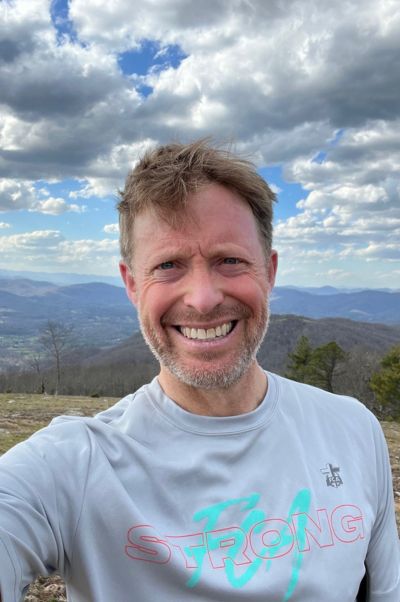 A person smiles for a selfie on a mountainous landscape with scattered clouds and distant hills in the background.