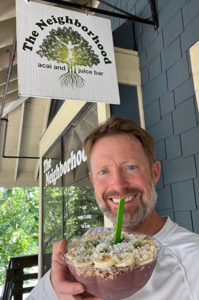 A person holding an acai bowl stands under a sign reading "The Neighborhood Acai and Juice Bar." Background includes greenery and building exterior.