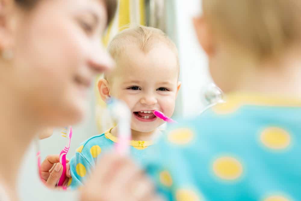 A child and a person are brushing teeth together, smiling in a mirror. Both wear blue outfits with yellow dots. Bright, cheerful scene.