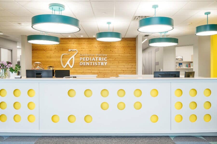 Bright dental office reception with modern lighting and decor. "Pediatric Dentistry" on wooden wall. Yellow accents adorn the white counter.