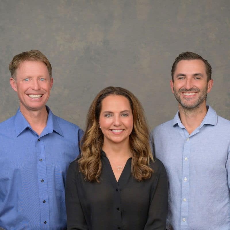 Three people standing and smiling, dressed in casual button-up shirts against a plain, gray studio backdrop.