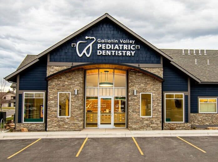 A modern building with navy siding and stone accents houses Gallatin Valley Pediatric Dentistry. It's situated in a spacious parking lot under a cloudy sky.