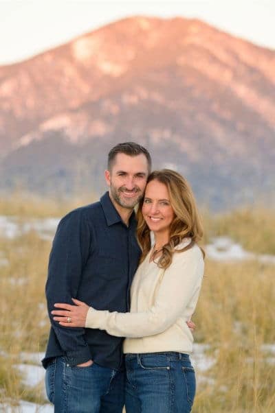 A person in a dark shirt embraces another in light clothing, standing in a grassy field with a mountain in the background.