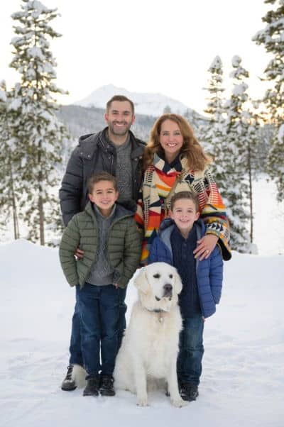 A family of four and a dog pose in a snowy forest with mountains in the background, all wearing winter clothing.