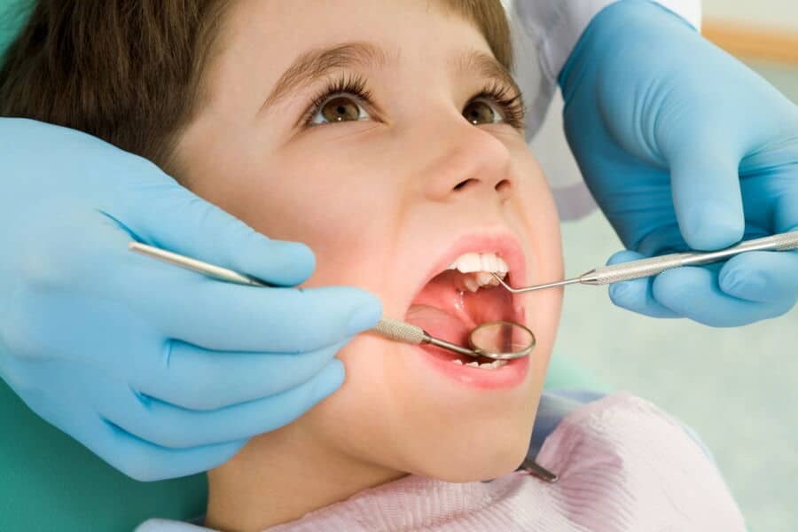 A child receives a dental check-up. A person wearing blue gloves uses dental instruments to examine the child's open mouth.