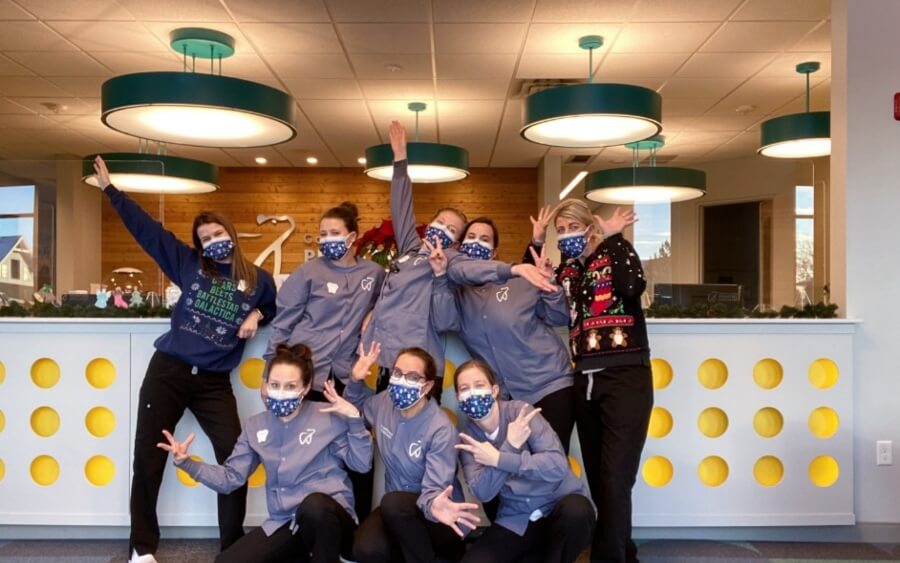 Eight people pose enthusiastically indoors, wearing matching blue jackets and masks. The background features a modern interior with circular ceiling lights.