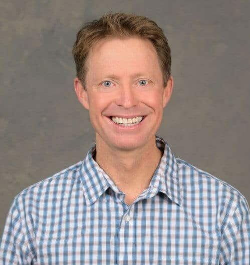 A person wearing a blue and white checkered shirt smiles against a plain, textured background.