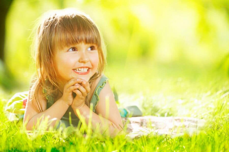 A smiling child lies on vibrant green grass, surrounded by sunlight and soft focus foliage, creating a joyful and serene outdoor scene.