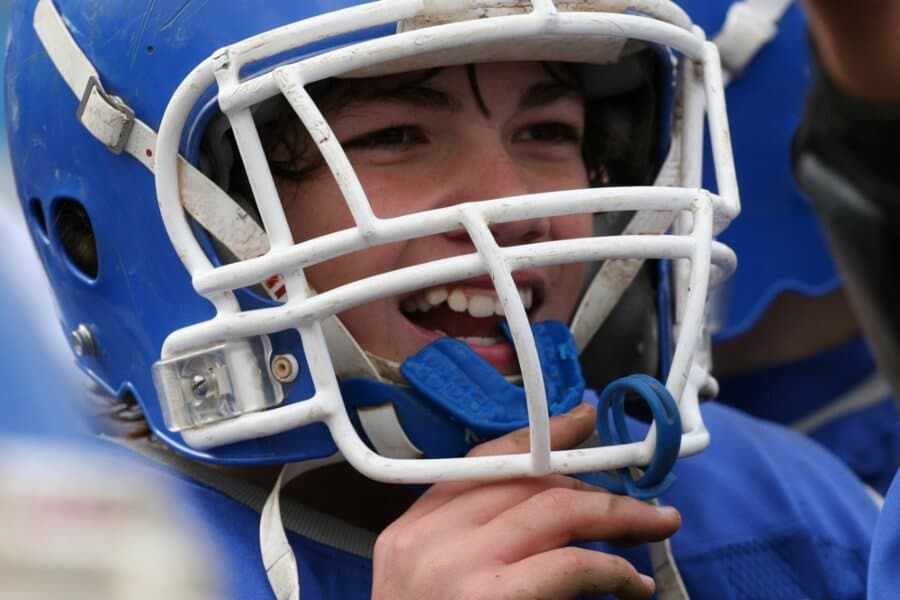 A person wearing a blue football helmet and jersey smiles, looking excited. The scene captures a moment of enthusiasm and teamwork in sports.