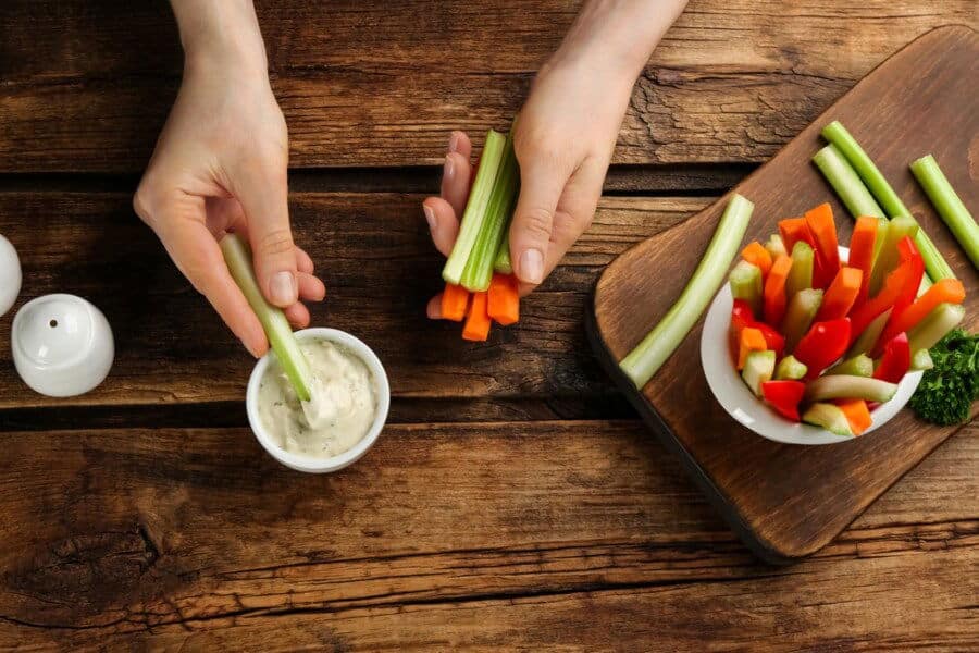 A person dips celery into a white sauce. A wooden board displays colorful vegetable sticks, including peppers and carrots, on a rustic table.