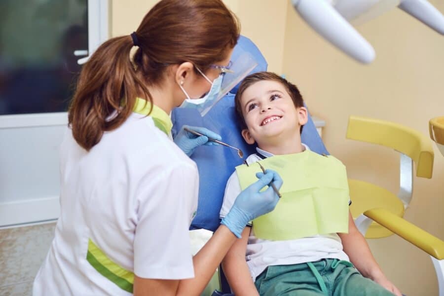 A child in a dental chair smiles at a person wearing gloves and a mask, holding dental tools in a brightly lit clinic.