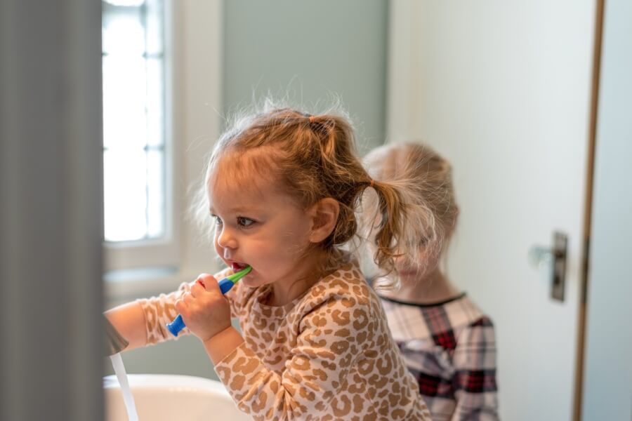 Two young children brushing teeth in a bathroom, one in focus, the other blurred. A window is visible in the background.