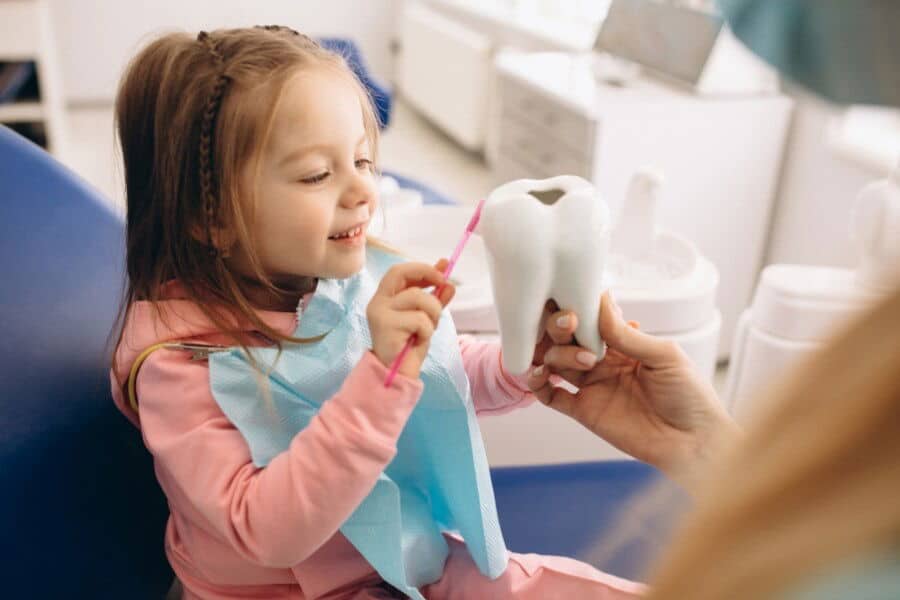 A child is brushing a large tooth model with a pink toothbrush, while a person holds it, in a dental office setting.