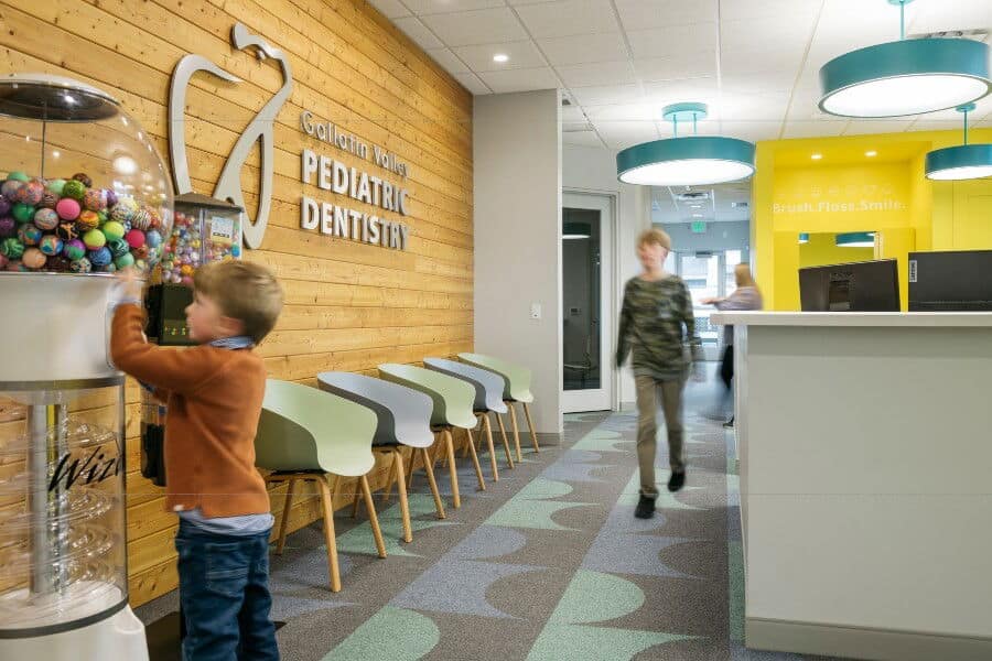 A dental office with playful decor, featuring children near a gumball machine and a bright waiting area with colorful chairs and signage.