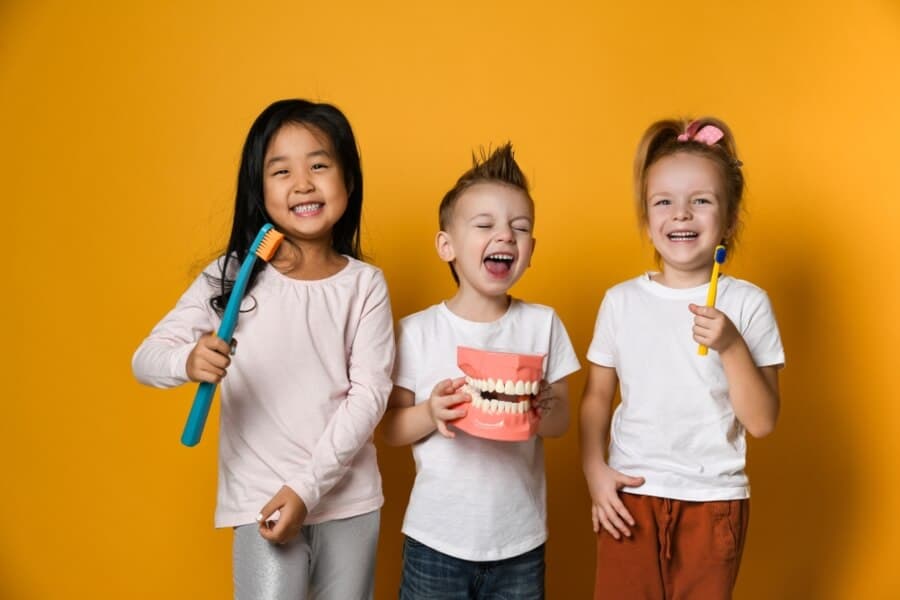 Three children smiling against a yellow background. They hold toothbrushes and a dental model, showcasing healthy dental habits.