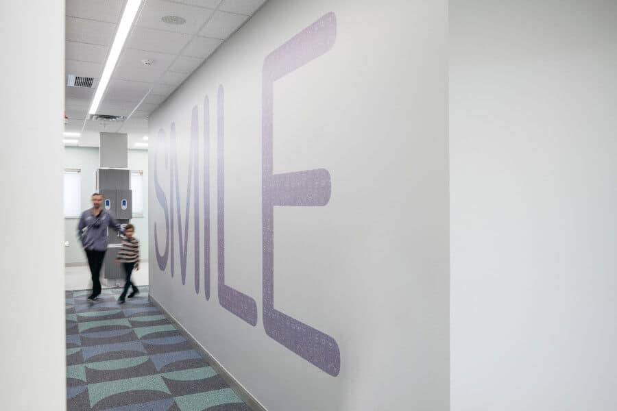 A person and child walk down a hallway with a large "SMILE" sign on the wall. The floor has a patterned design.