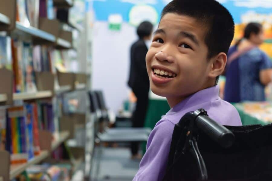 A smiling child in a wheelchair, wearing a purple shirt, looks back in a library setting with blurred books and people around.