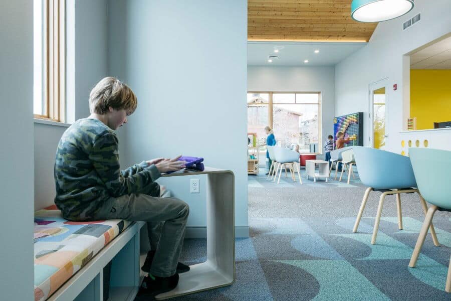 A child seated on a colorful bench uses a tablet in a bright room with modern furniture and another child in background.