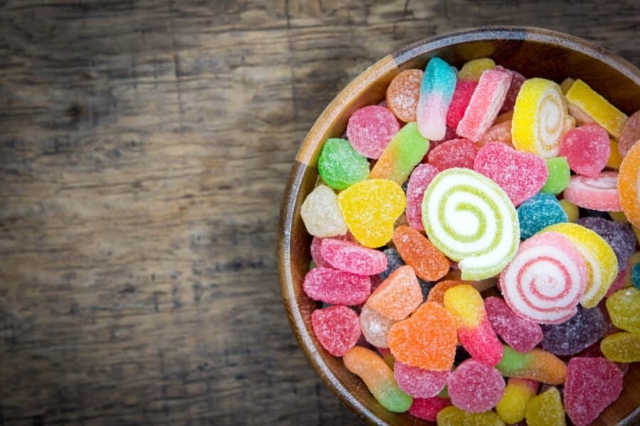 A wooden bowl filled with colorful gummy candies and jelly sweets on a rustic wooden surface, featuring spiraled designs and sugar-coated textures.