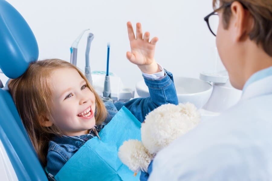 A child in a dental chair smiling and holding a teddy bear, with a person in a white coat in a dental office.