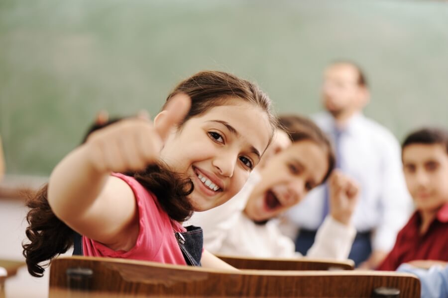 A group of children in a classroom setting, smiling and giving a thumbs-up, with a person in the background near a chalkboard.